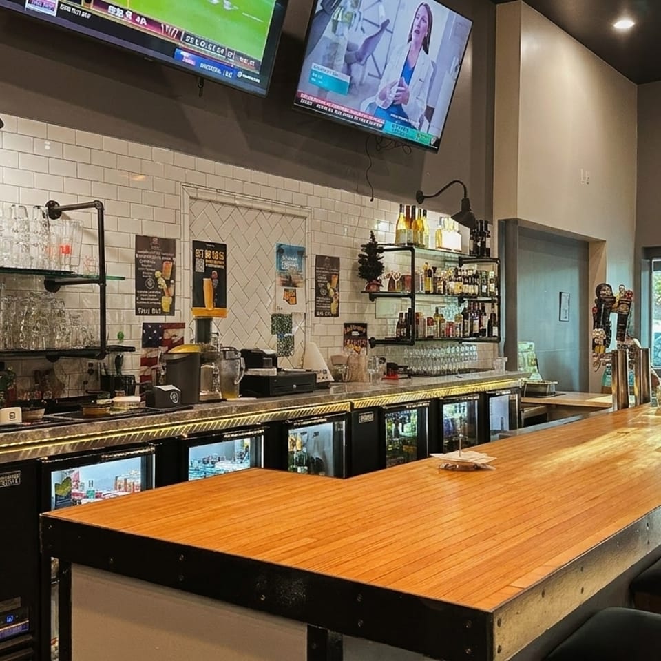 A well-lit bar area with a long wooden counter, black stools, and TVs mounted above.