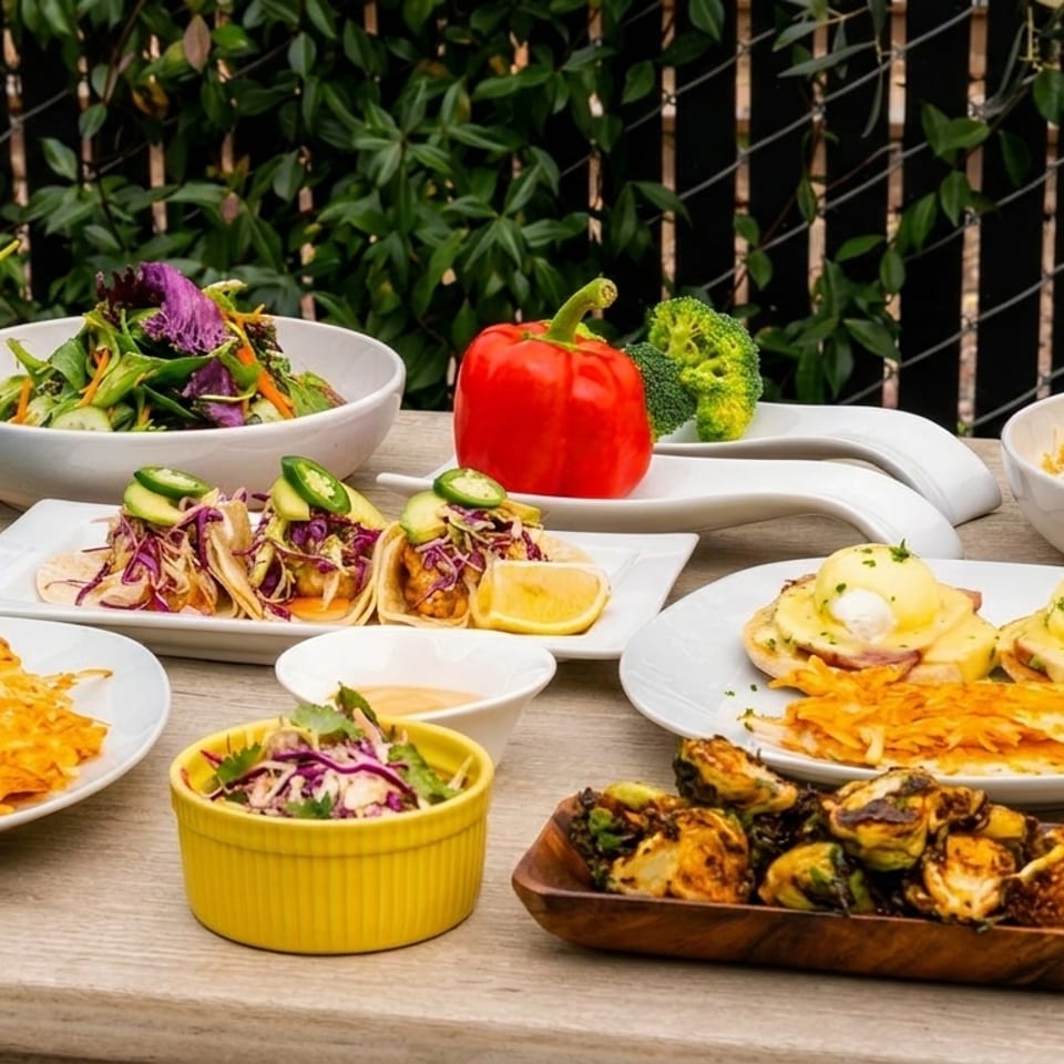 Outdoor table laden with various dishes including tacos, eggs Benedict, hash browns, salads, and roasted vegetables.