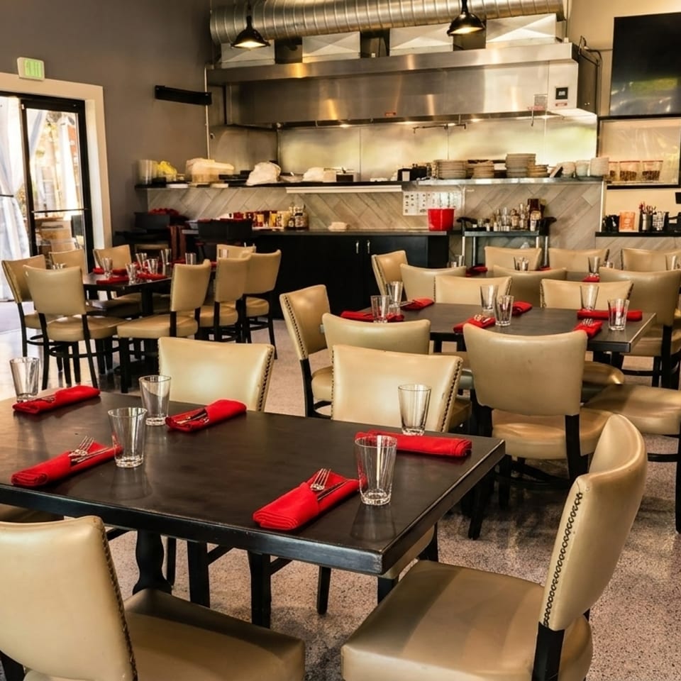 Empty restaurant dining room with tables set with red napkins and an open kitchen in the background.